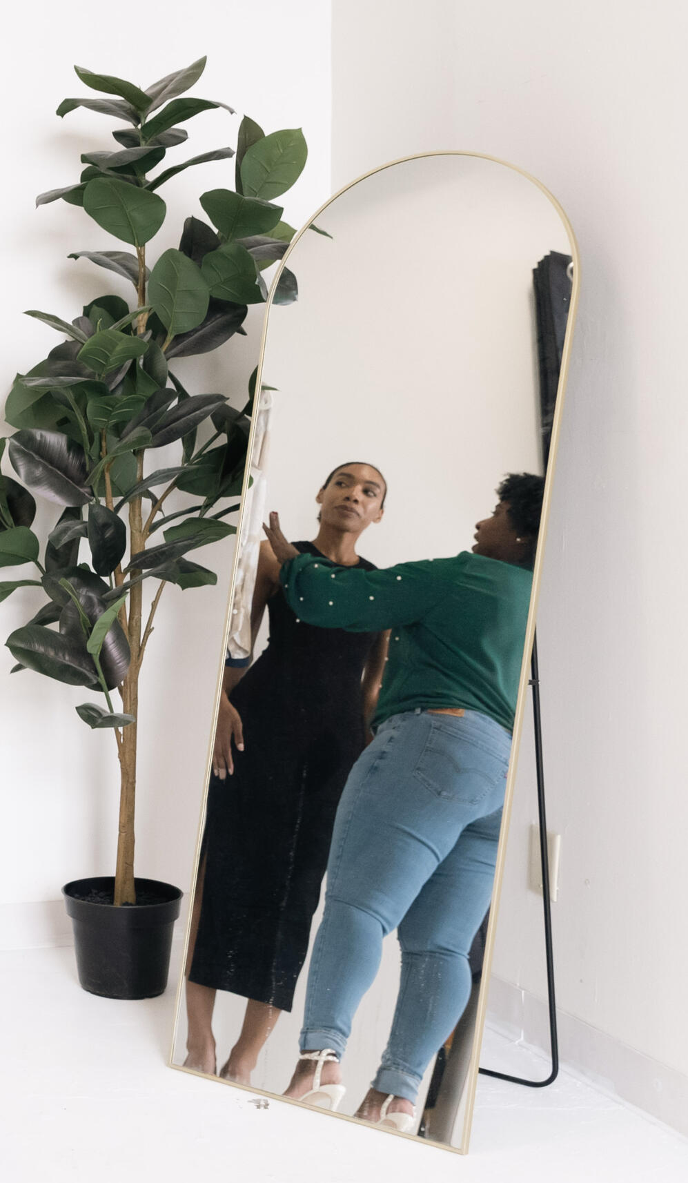 iconic-styling-session-client-fitting-gold-mirror.jpg Personal stylist adjusting a client’s outfit in a mirror beside a clothing rack and plant.