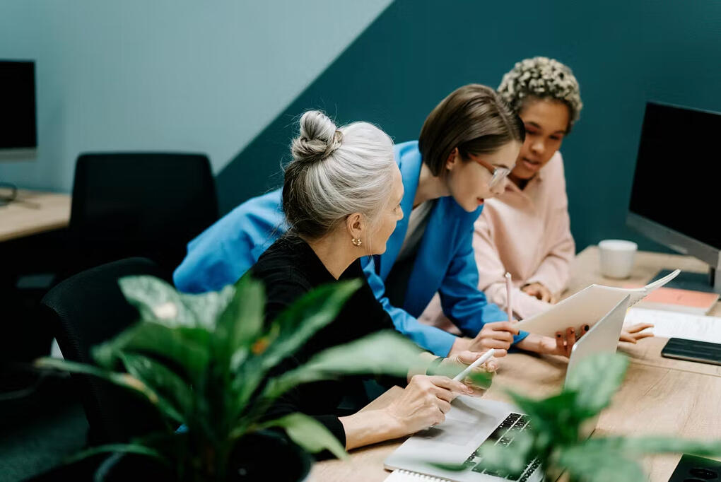 Three women collaborating at a conference table, reviewing notes and a laptop in a modern workspace.